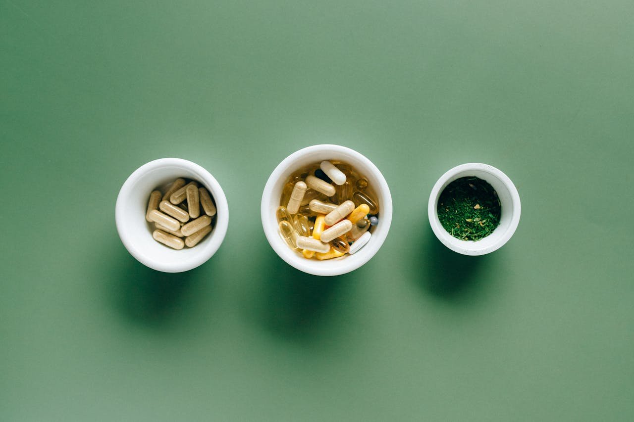 Top view of bowls with capsules, herbs, and supplements on green surface for healthcare concept.